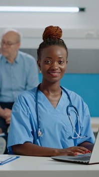 Portrait of nurse typing on laptop keyboard in medical office at facility. Assistant woman with uniform sitting at desk using gadget with technology. Doctor and old patient in background