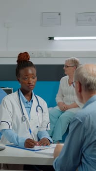 Young medical assistant giving x ray to doctor sitting at desk with elder patient for scan results analysis in office. Medic holding radiography while old man looking at healthcare diagnosis