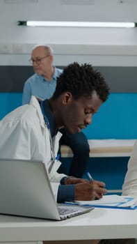 Healthcare specialist writing medical information at appointment with senior patient. Doctor in hospital room with old woman at desk while nurse and elder man sitting in background