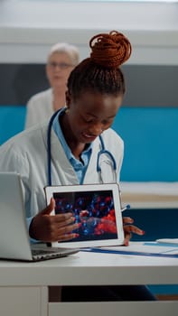 Black doctor holding virus animation on tablet showing disease cells to elder patient at desk. African american medic explaining coronavirus bacteria using modern technology on device.