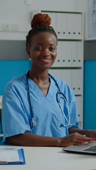 Portrait of healthcare nurse typing on laptop keyboard for medical consultation and appointment while sitting at desk. Black woman smiling with assistant uniform and stethoscope