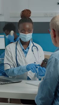 Black professional doctor showing bottle of pills to elder patient against sickness in medical office with glass protection. African american medic giving advice, and treatment to senior man