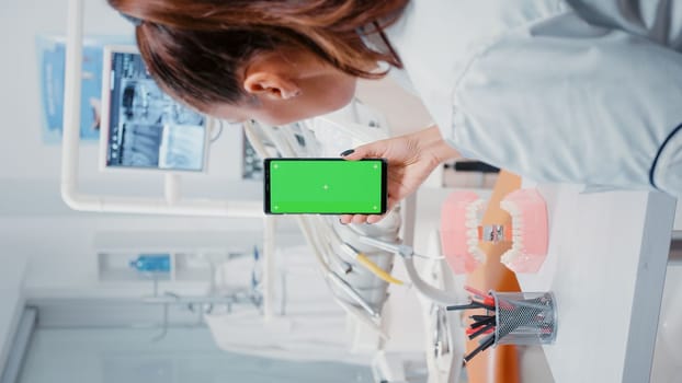 Vertical video: Woman vertically holding mobile phone with green screen in dental cabinet. Dentist looking at isolated mockup background with chroma key on display for dentistry.