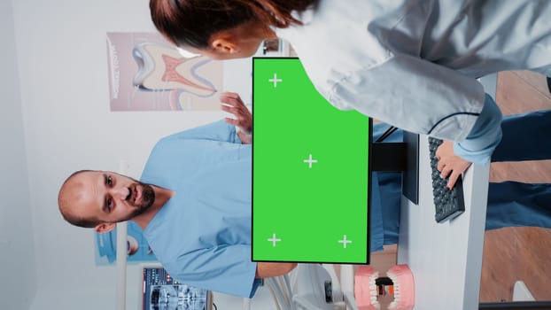 Vertical video: Dentist working with horizontal green screen on computer while talking to assistant in dental office. Orthodontist using chroma key and mockup template for oral care and dentistry