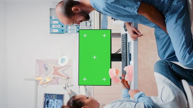 Vertical video: Team of specialists working with green screen on monitor in dental office. Dentist and man assistant using chroma key with mockup template and isolated background for oral care