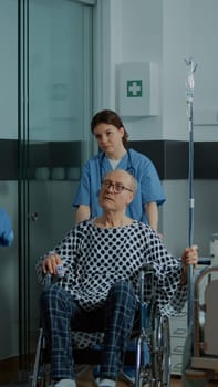 African american doctor taking sick patient in wheelchair from hospital ward to surgery room. Old man with IV drip bag leaving bed in unit with modern technology medical equipment