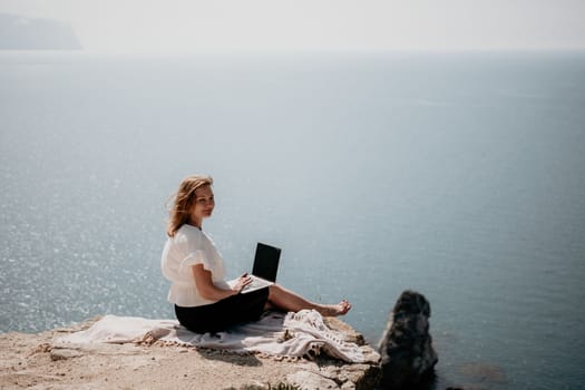 Successful business woman in yellow hat working on laptop by the sea. Pretty lady typing on computer at summer day outdoors. Freelance, travel and holidays concept.