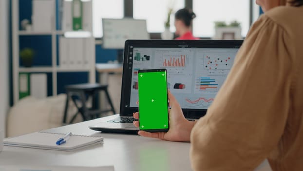 Close up of woman vertically holding smartphone with green screen in business office. Person looking at mobile phone with isolated mock up background and chroma key template on display.