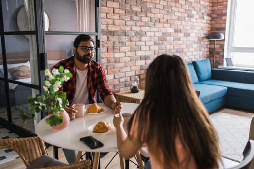 Young diverse loving couple eating croissant and talks together at home in breakfast time. Communication and relationship