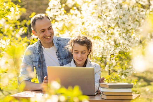 A girl and a young father are sitting at a laptop and studying in a flowered garden. Against the background of green grass and flowering trees. Remotely buy items in a store.