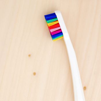 White toothbrush with multicolored bristles on a wooden table. Bristles in all colors of the rainbow. Rainbow toothbrush with white knob. Fashionable oral care.