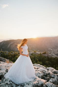 Bride in a white puffy dress stands on a rock at sunset. High quality photo