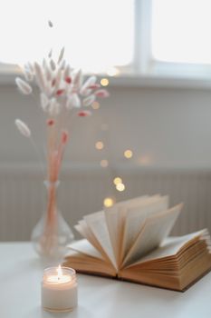 A minimalistic composition in the Scandinavian style with book, dried flowers in a vase and candle by window. Cozy home