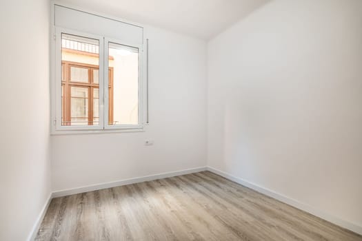 An empty bright room with a window in which the windows of a nearby house are visible. External metal shutters are built into the window to darken the room during daylight hours