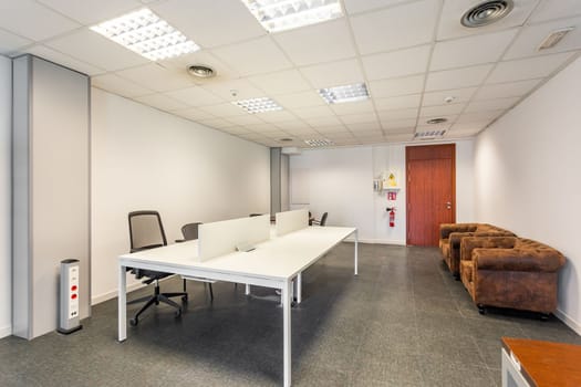 Large conference room with tiled floor and bright lamps on the unfashionable ceiling. Along the walls are outdated armchairs with worn upholstery