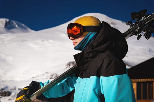 The skier holds a pair of skis and looks at the snow-capped mountains to the side. A guy with skis on his shoulder stands in the mountains covered with snow. Man with winter gear having fun.