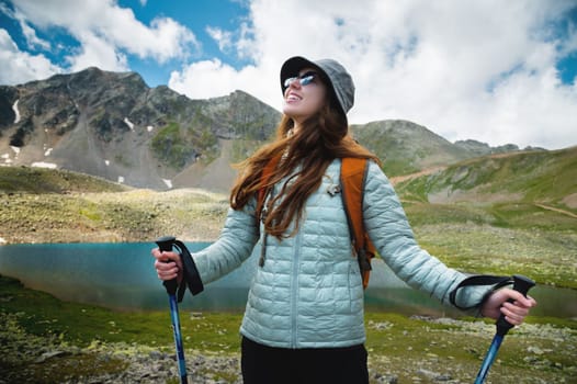 Portrait of a tourist with a backpack, front view. A young girl leads an active lifestyle, walks along a mountain path with trekking poles, mountain peaks and a lake in the background.
