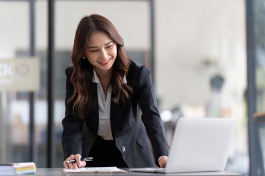 Young business woman standing in office writing work using laptop and take note.
