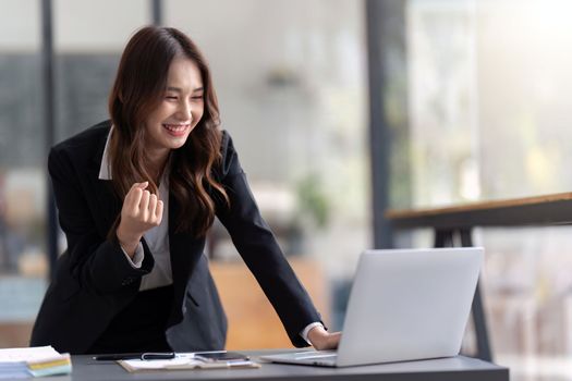 Asian Happy businesswoman sitting at desk in office feel euphoric win online, Business woman celebrating a triumph with arms up.