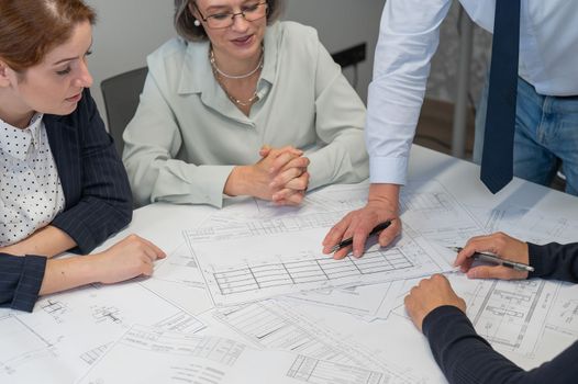 A caucasian man is standing making changes to a drawing, three colleagues are sitting at a table and listening to him