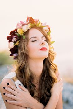 Smiling bride in a flower wreath hugging herself by the shoulders, raising her head to the sun. High quality photo