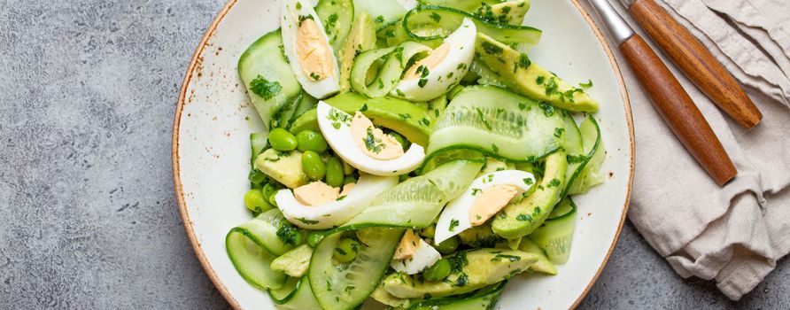 Healthy green avocado salad bowl with boiled eggs, sliced cucumbers, edamame beans, olive oil and herbs on ceramic plate top view on grey stone rustic table background