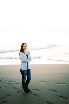 Young woman in a sweater and jeans stands on the beach with her arms crossed over her chest. High quality photo