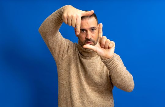 Handsome bearded happy male looks through a frame formed by his hands. An attractive man makes a frame with his fingers, isolated on a blue background with copy space for text. People concept