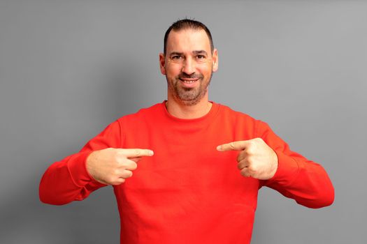 Bearded hispanic man wearing a red sweater pointing to himself proudly isolated on gray background. He is satisfied with his work, he feels effective and competitive