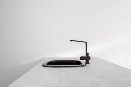 Black metal kitchen sink with trendy faucet sits on large white stone kitchen island on light background. Concept of kitchen after renovation or in new building. New plumbing.