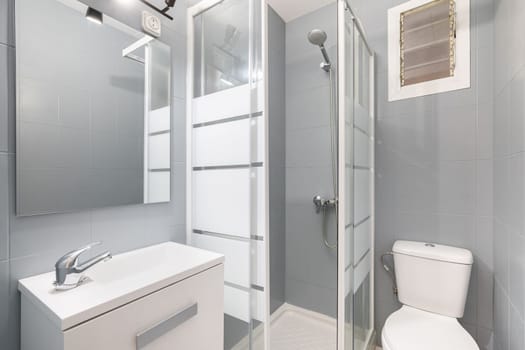 Compact modern bathroom in gray and white tones with shower with glass doors, toilet bowl and sink with cabinet for storing things. Concept of a toilet design idea in a compact apartment.