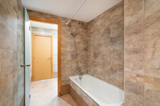Bathroom with marble tile walls with abstract multicolored strokes. It is convenient to take relaxing procedures in a white bath. Doorway reveals corridor with door on opposite side