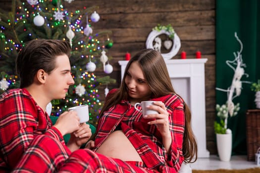 A couple expecting a baby sits on a gray couch against a backdrop of a dressed Christmas tree with lights. The man is holding the woman on his lap, both are holding white cups and looking into each other's eyes.