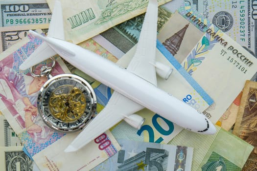 Pocket watch, airplane sitting on arrangement of international currencies including euro, ruble USD. Concept of high cost of travel. High quality photo