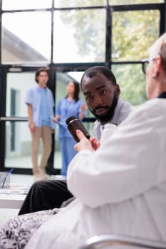 Elderly physician showing painkiller bottle to african american patient while explaining medication treatment during checkup visit appointment in hospital lobby. Medicine service and concept