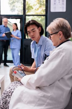 Elderly cardiologist measuring asian patient hypertension and blood pressure to do heart exam with tonometer in hospital waiting area. Doctor consulting young adult during checkup visit consultation