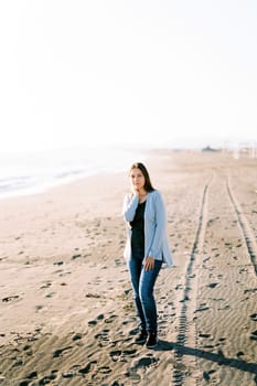 Young woman in a sweater stands on a sandy beach by the sea. High quality photo