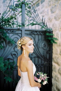 Bride with a bouquet stands sideways near a stone house. High quality photo
