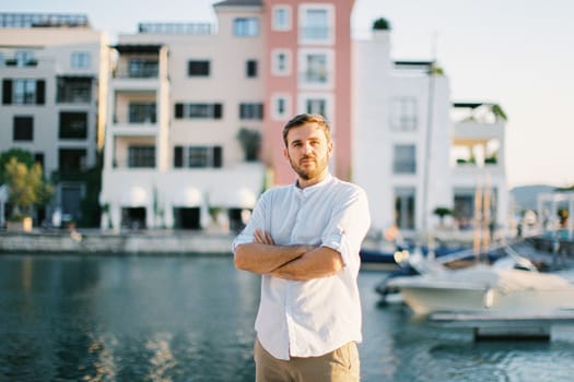 Young man with his arms crossed on his chest stands against the backdrop of moored yachts. High quality photo