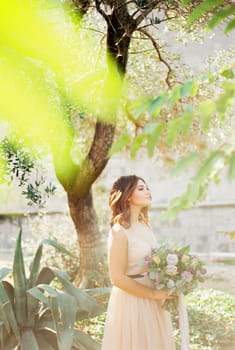 Bride with a bouquet stands near a green tree and looks away. High quality photo