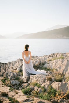 Bride in a white dress stands on a rocky seashore and looks at the mountains. Back view. High quality photo