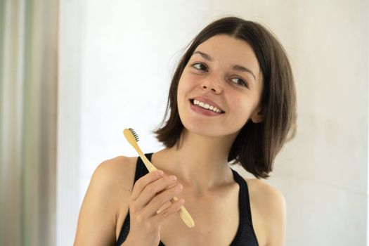 A young smiling girl brushes her teeth with toothpaste in the bathroom by the mirror in the morning, a woman takes good care of the health of tooth enamel and gums.