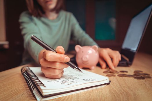 A young girl works at a laptop, writes a business development plan in a notebook, develops a strategy and makes calculations of earned funds. A woman puts her savings in a piggy bank.