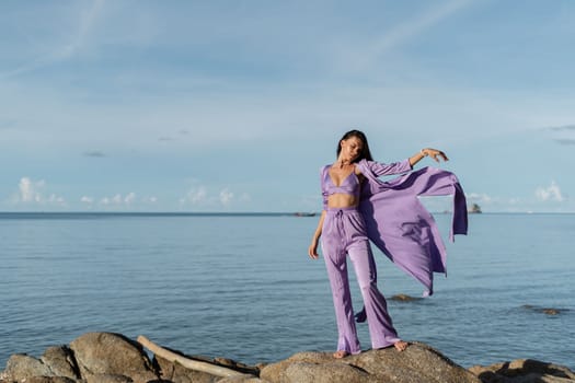 Young beautiful woman in a romantic mood, lilac silk clothes, on the beach against the backdrop of the sea and stones at sunset