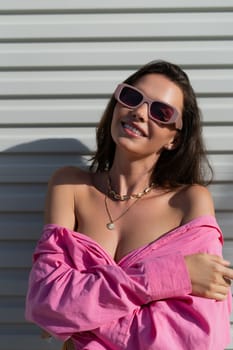 Young beautiful brunette in a pink shirt, neck jewelry, necklace, trendy sunglasses on the background of a light garage door fence