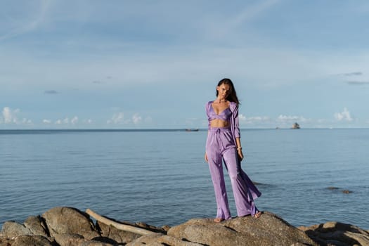 Young beautiful woman in a romantic mood, lilac silk clothes, on the beach against the backdrop of the sea and stones at sunset