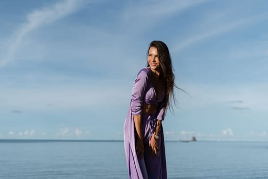 A young beautiful woman in a romantic mood, lilac silk clothes, on the beach against the backdrop of the sea and stones at sunset, laughs, smiles with a snow-white smile