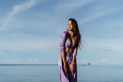 Young beautiful woman in a romantic mood, lilac silk clothes, on the beach against the backdrop of the sea and stones at sunset