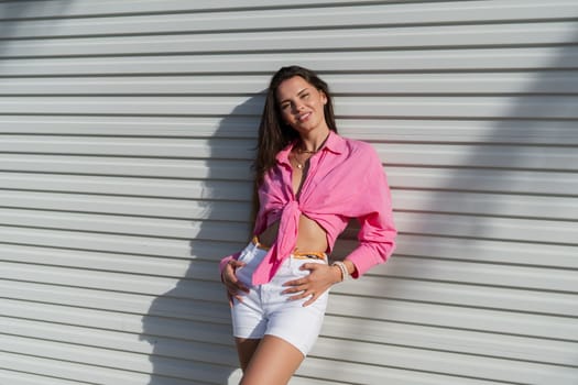 Young beautiful brunette woman in a pink shirt and white denim shorts against the background of a light garage door fence