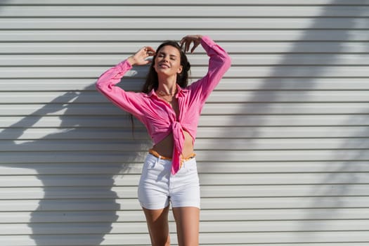 Young beautiful brunette woman in a pink shirt and white denim shorts against the background of a light garage door fence